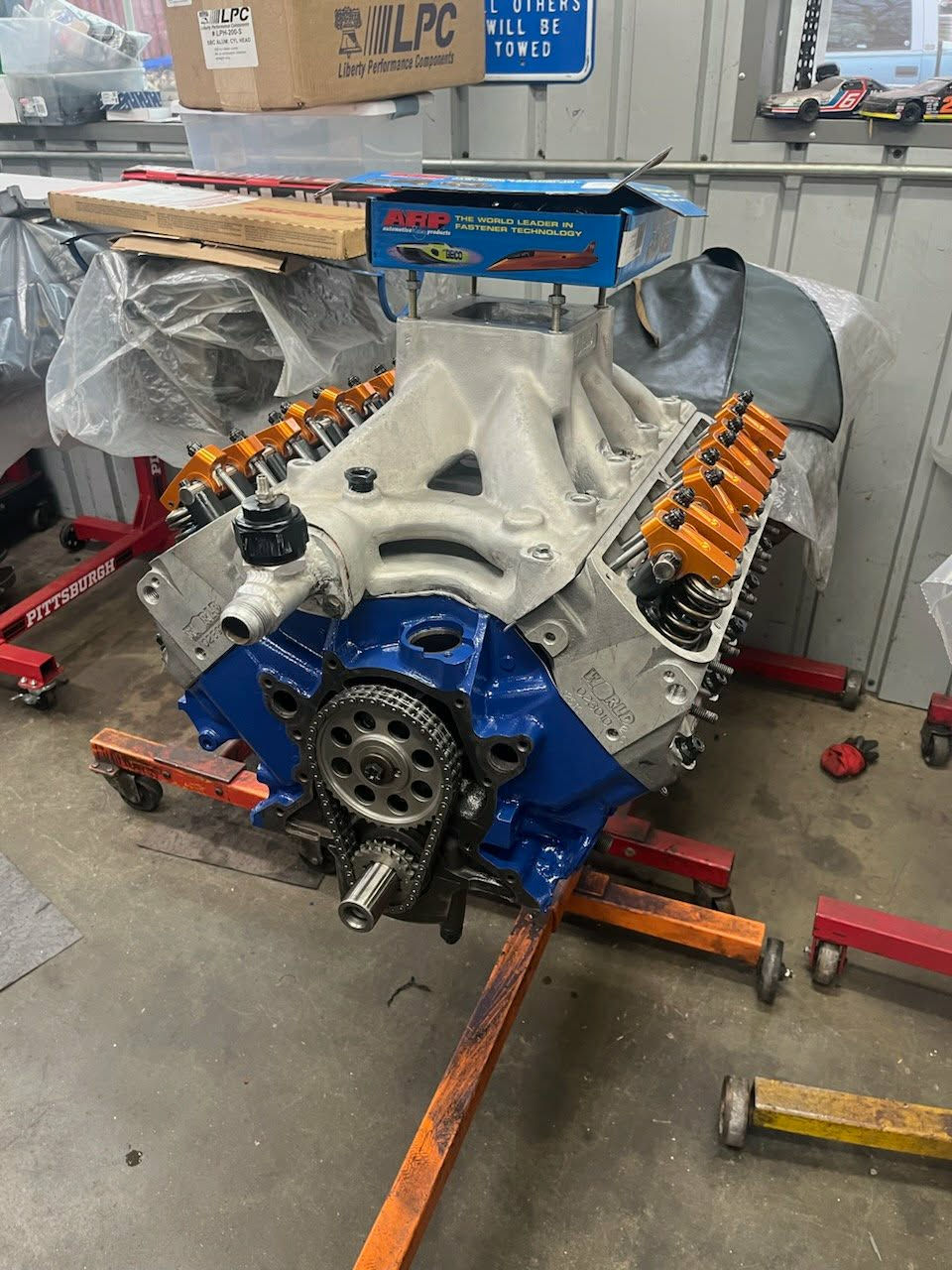 Custom blue engine block assembly with aluminum heads, orange roller rocker arms, and high-rise intake manifold on an engine stand."