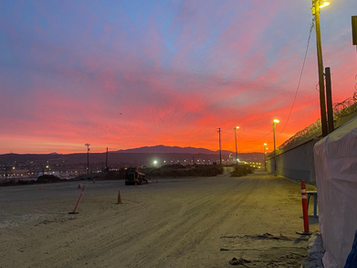 A dirt path at dawn lined with floodlights and a tall border fence on the right. The sky glows in dramatic hues of pink, orange, and blue as the sun rises behind distant mountains. Tarps and cones mark the area, evoking both beauty and the stark reality of the border zone.