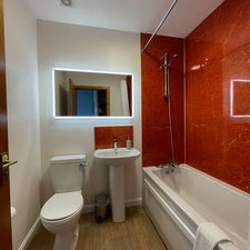 Bathroom with red wet wall surrounding a big bath tub and a white sink, with LED mirror lit above, bin and toilet.