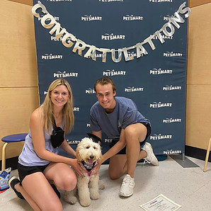 Couple kneels with their white goldendoodle, who is sitting & wearing a graduation cap, in front of a PetSmart backdrop with sign that says "Congratulations".