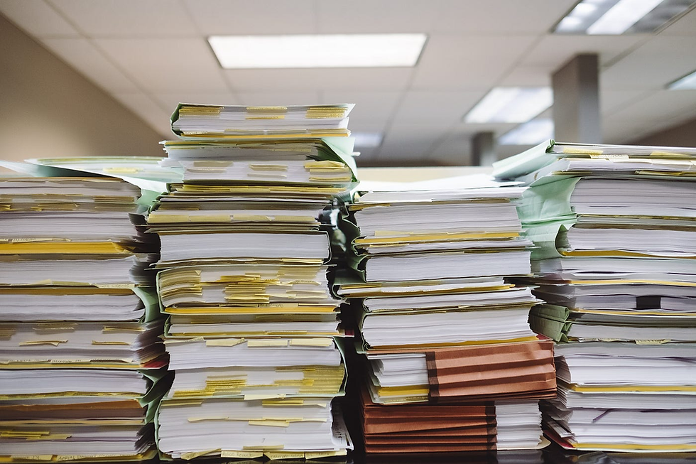 Stacks of paper files with yellow tabs on a desk in an office. Ceiling lights illuminate the scene, suggesting a busy, organized environment.