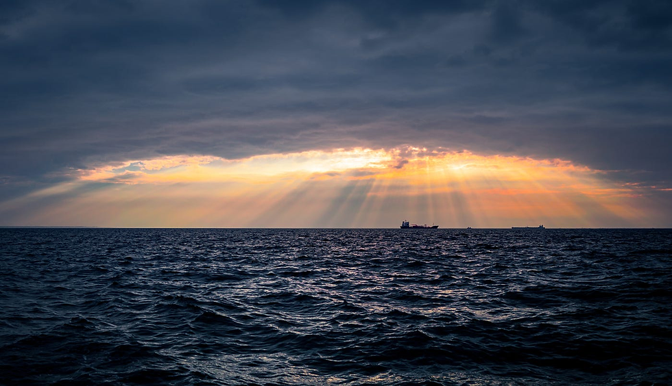 Sunset over ocean with dark waves; rays of sunlight break through clouds, illuminating a silhouetted ship on the horizon. Calm, dramatic scene.