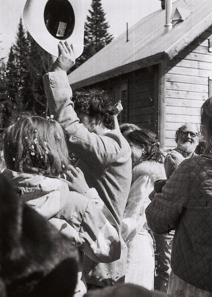Photo de mariage noir et blanc sur film, invités en liesse lançant un chapeau devant une maison de bois