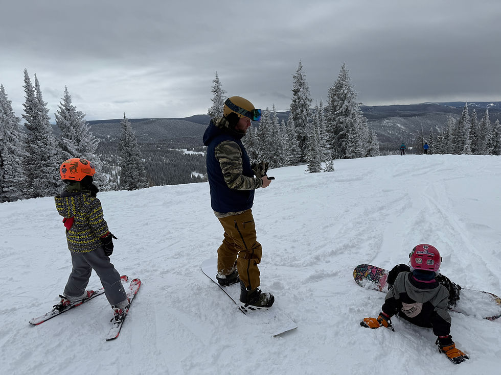 A snowboarder and two children on skis pause on a snowy mountain. Overcast sky, snow-covered trees, bright helmets, and playful mood.
