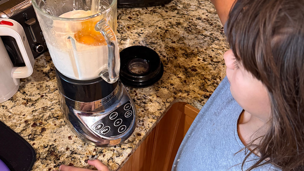 Child pouring liquid into a blender in a kitchen with granite countertop. Wearing a gray shirt and red pants, focused on the task.
