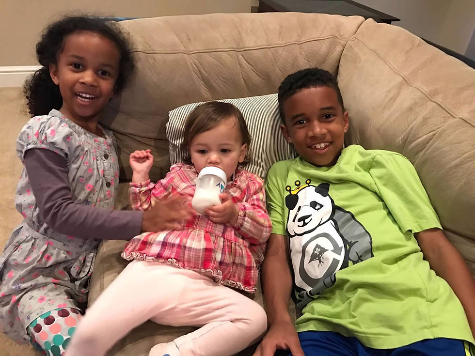 Three smiling children on a beige couch: one in a floral dress, another in plaid drinking from a bottle, and a third in a green panda shirt.