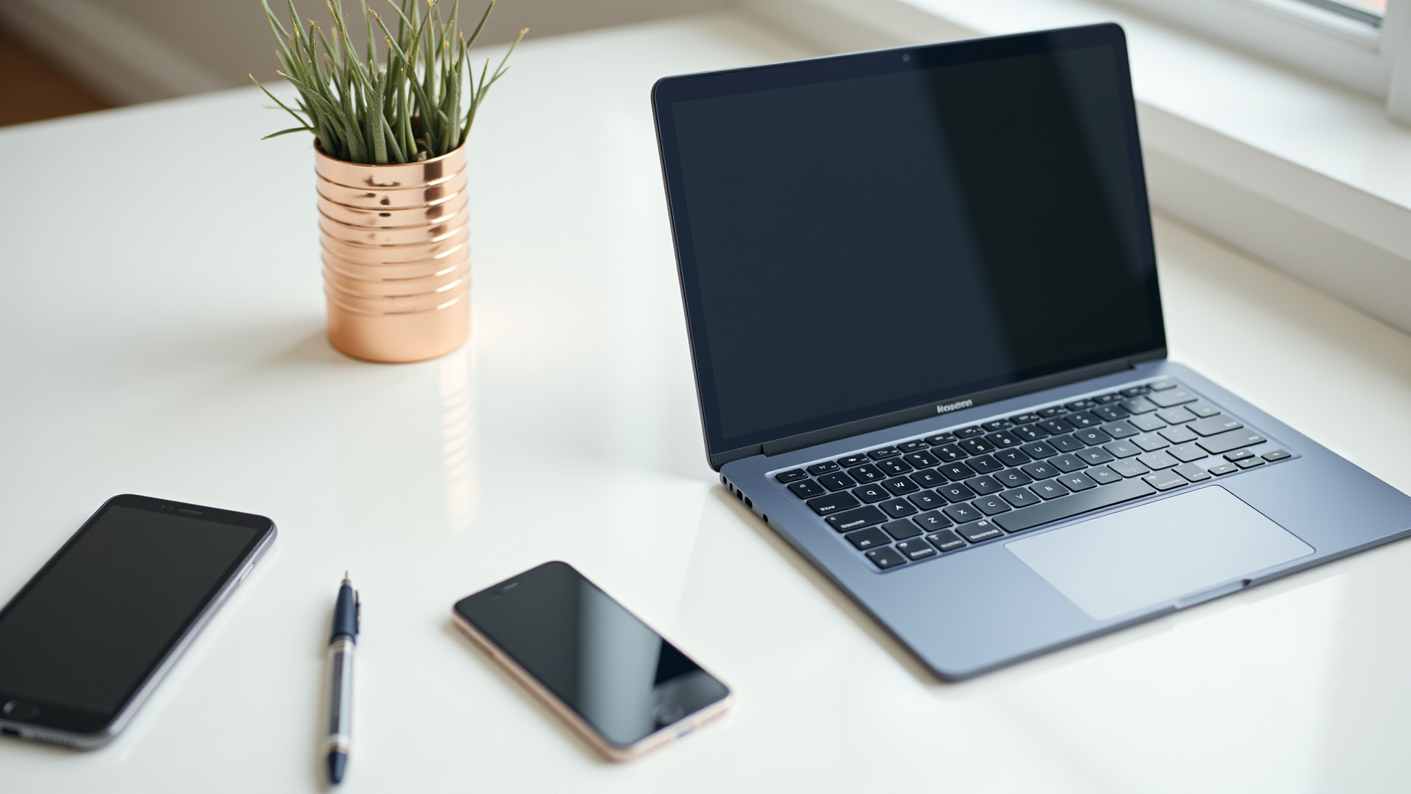 Laptop, dois smartphones, caneta e planta sobre mesa branca.