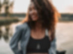 A smiling woman standing near a lake. She’s wearing a black Unalome Journey sports bra under a jean jacket.
