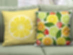 Two Citrus Burst throw pillows on green couch. Front side orange, lime, lemon, strawberries. Back side one big lemon slice.