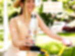 A happy young woman wearing a sunhat is riding her bike and holding a Happy Flowers water bottle.