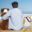 Thumbnail: Man sitting on the beach with his arm around large dog. A Starfish Frolic tote bag is next to him filled with dog toys.