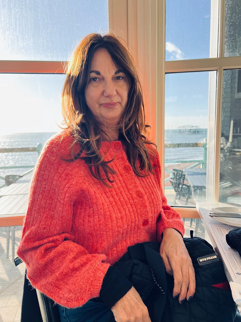 Mandy in a red sweater sits by a sunny window with a sea view, holding a black bag. Papers are on the table, creating a calm atmosphere.