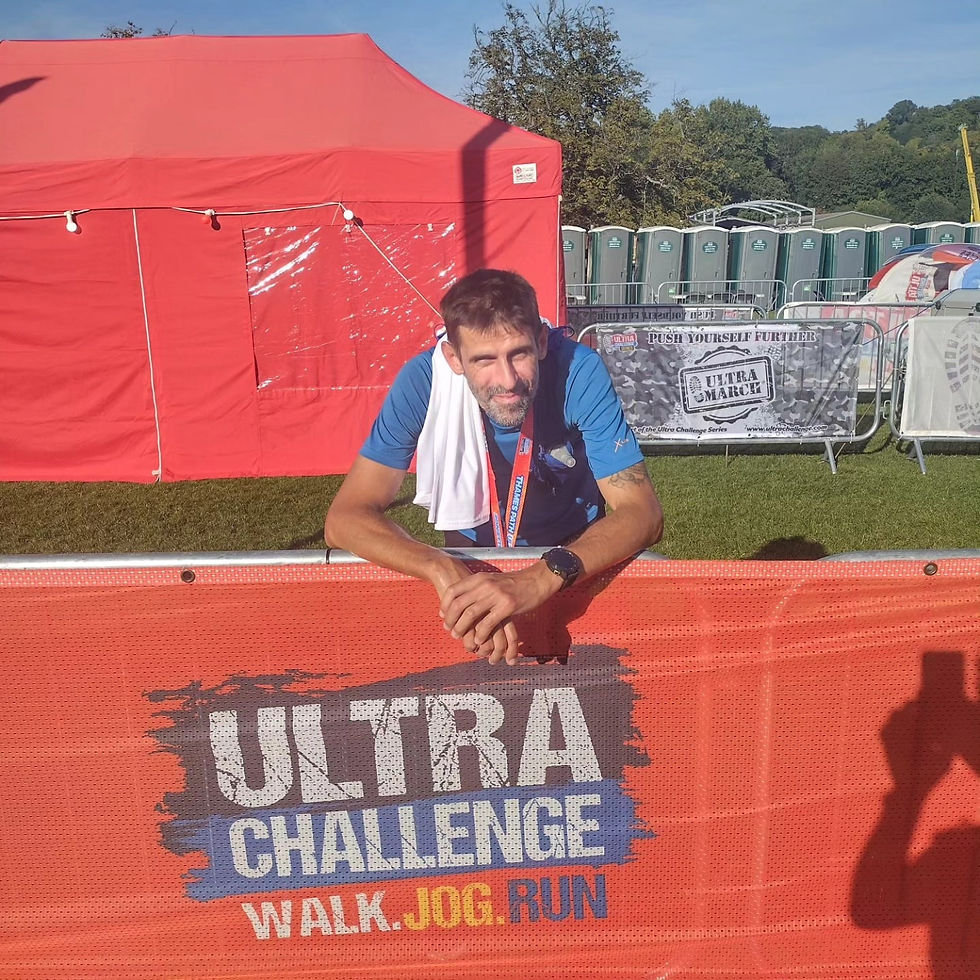 Michal leaning on an orange "Ultra Challenge" barrier, wearing a blue shirt. Red tent and green portable toilets in the sunny background.