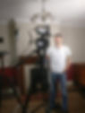 Man (Michal) in white t-shirt smiles, standing in front of a camera setup in a cozy room with wooden floor, soft lighting, and a chandelier.