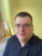 Bartek with glasses and a black shirt sits on a bed in a room with yellow walls and floral curtains, looking calm and relaxed.