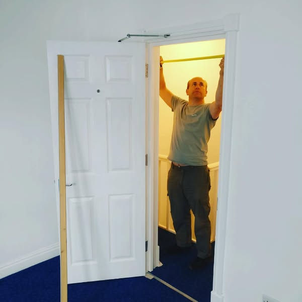 Man (Slawek) measuring door frame with tape. White walls, blue carpet, door ajar. Bright lighting from adjacent room. Focused expression.