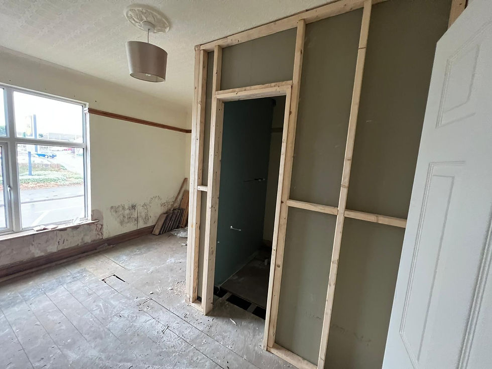 Interior view of a room undergoing renovation, showing exposed wooden studs framing a new wall and doorway, bare floorboards, a window with an outdoor view, and a partially removed wall section.