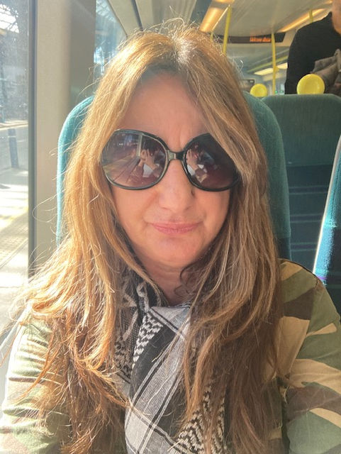 Mandy with long hair and sunglasses sits on a train in sunlight, wearing a patterned scarf. Reflection of passengers visible. Relaxed mood.