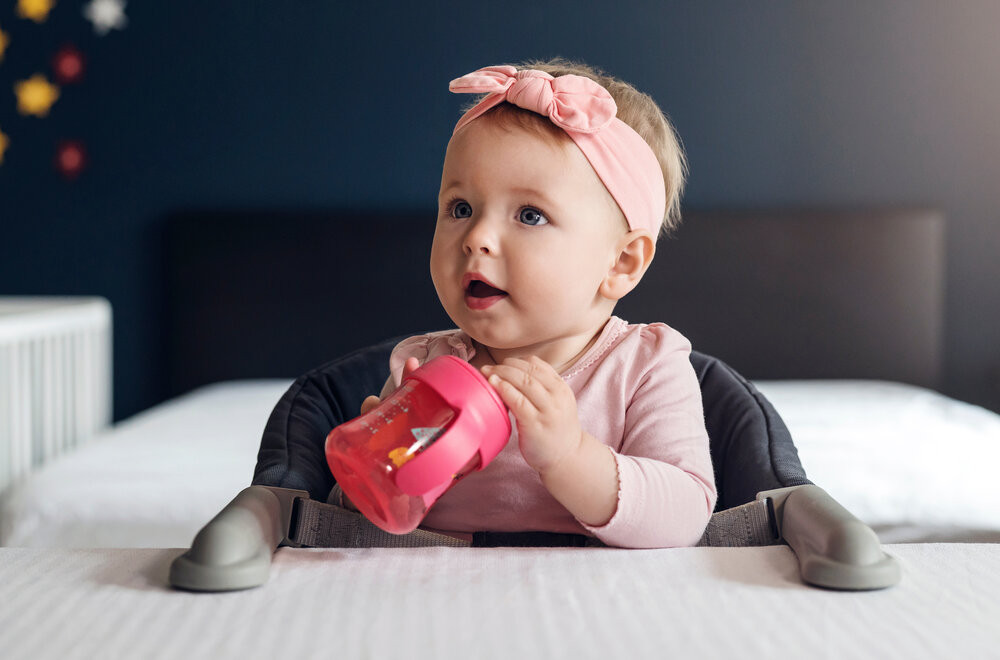 Baby with a pink bow sitting in a high chair holding a sippy cup