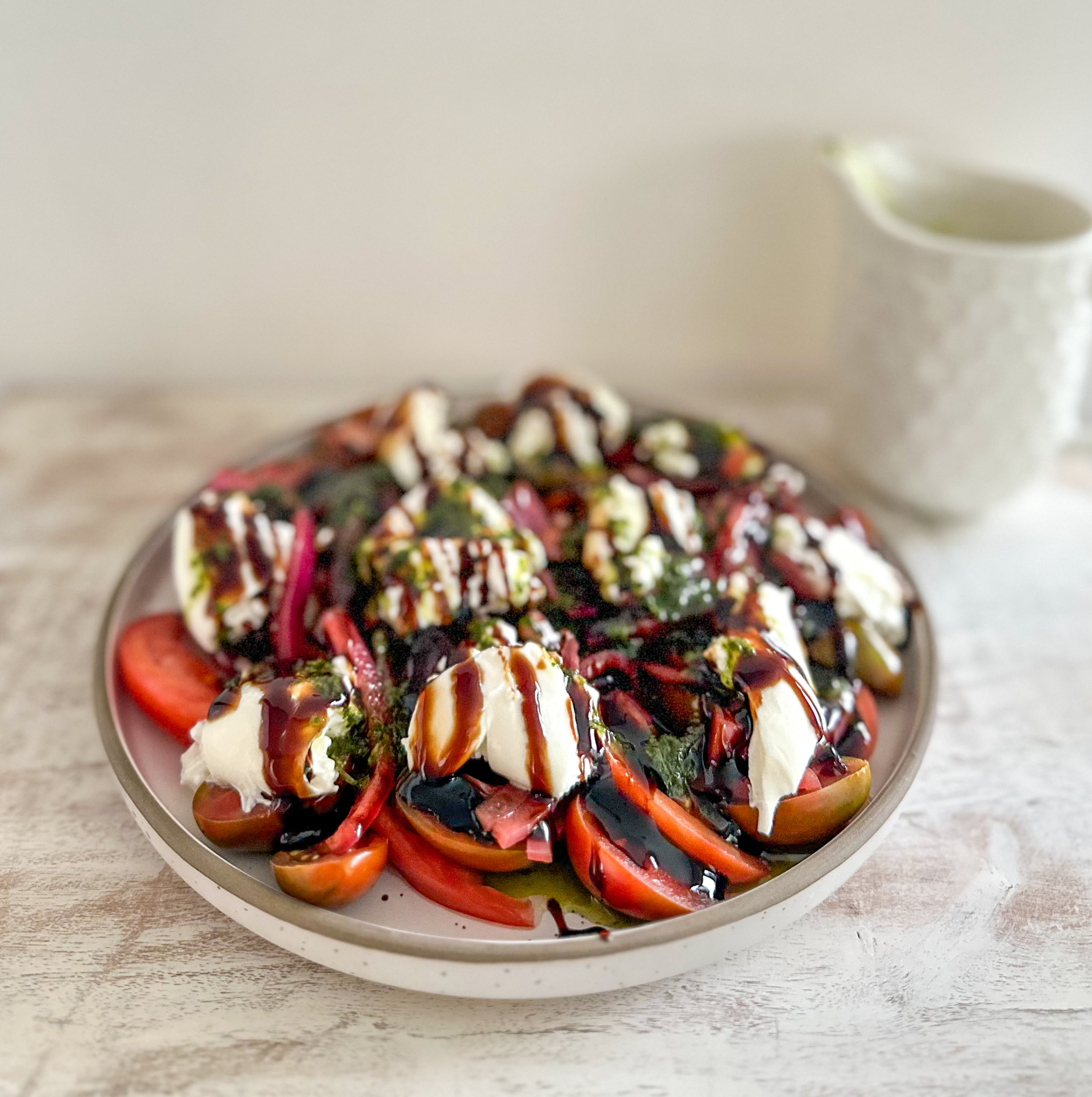 Salad with tomatoes, burrata, basil and balsamic drizzle on a plate beside a white mug on a soft-lit table
