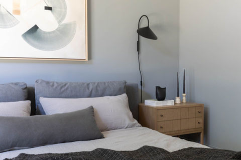Modern bedroom with gray upholstered bed, layered neutral bedding, black wall-mounted reading light, and light oak nightstand with stacked books and black decor.