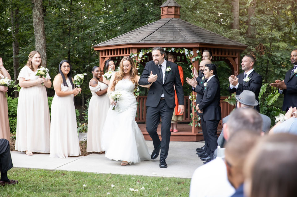 Newly married couple walking down the aisle smiling after outdoor gazebo wedding ceremony, surrounded by bridal party and guests