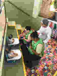 A imagem mostra duas cuidadoras dando de comer para dois bebes gêmeos, sentados em cadeirinhas especiais, no espaço tutti-frutti da Escola, enquanto a Nutricionista observa.