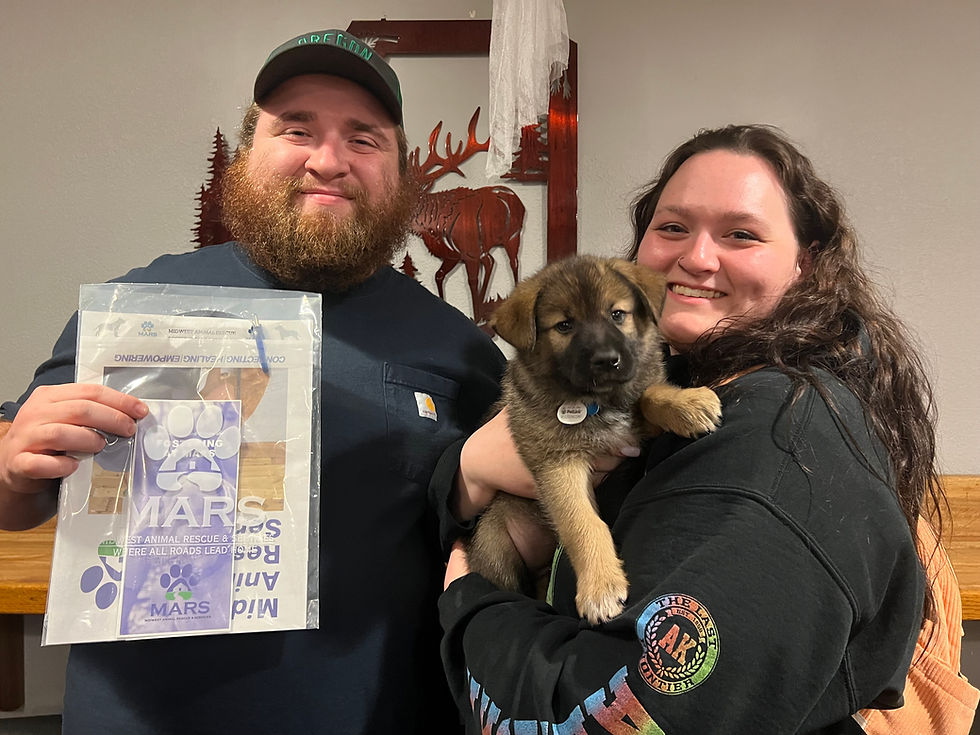 A man and woman smile while holding a young German Shepherd puppy at a MARS event. The man holds a MARS-branded bag featuring the "Where All Roads Lead Home" tagline.