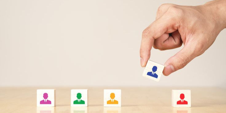 Hand placing a wooden block with a blue figure among blocks with colorful human icons on a wooden surface. Beige background, orderly setup.