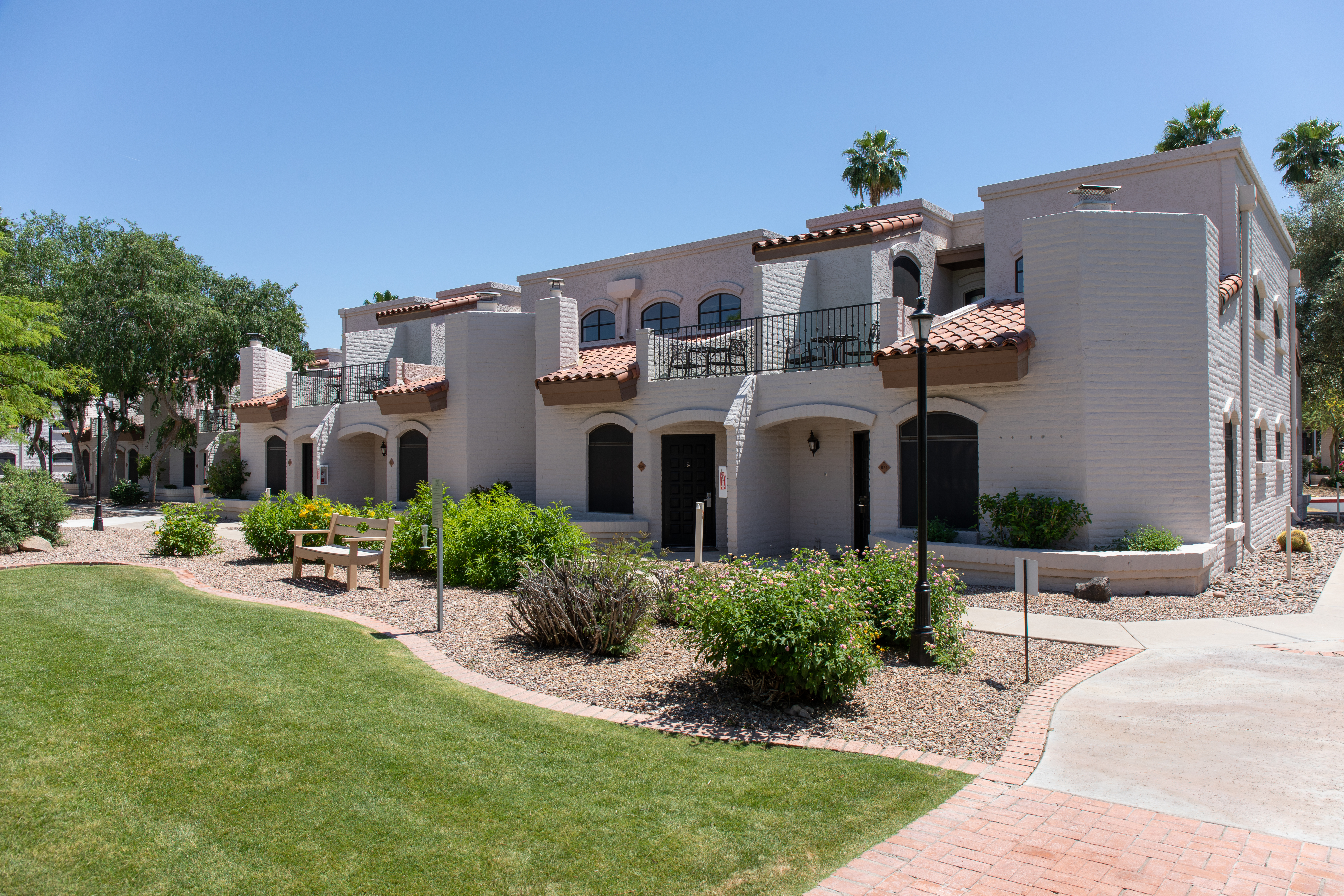 Cream stucco buildings with red tile roofs and arched doorways face a lawn with a bench, a lamp post and desert shrubs.