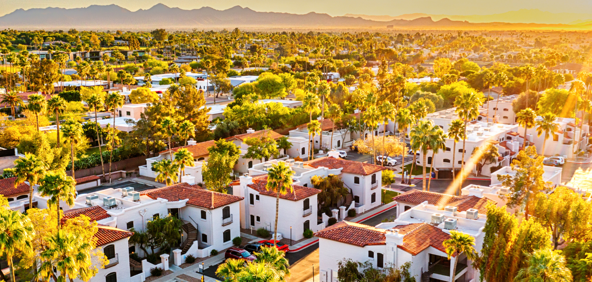 Golden light falls over dense trees, white stucco buildings with red clay roofs and a hazy mountain range in the distance.