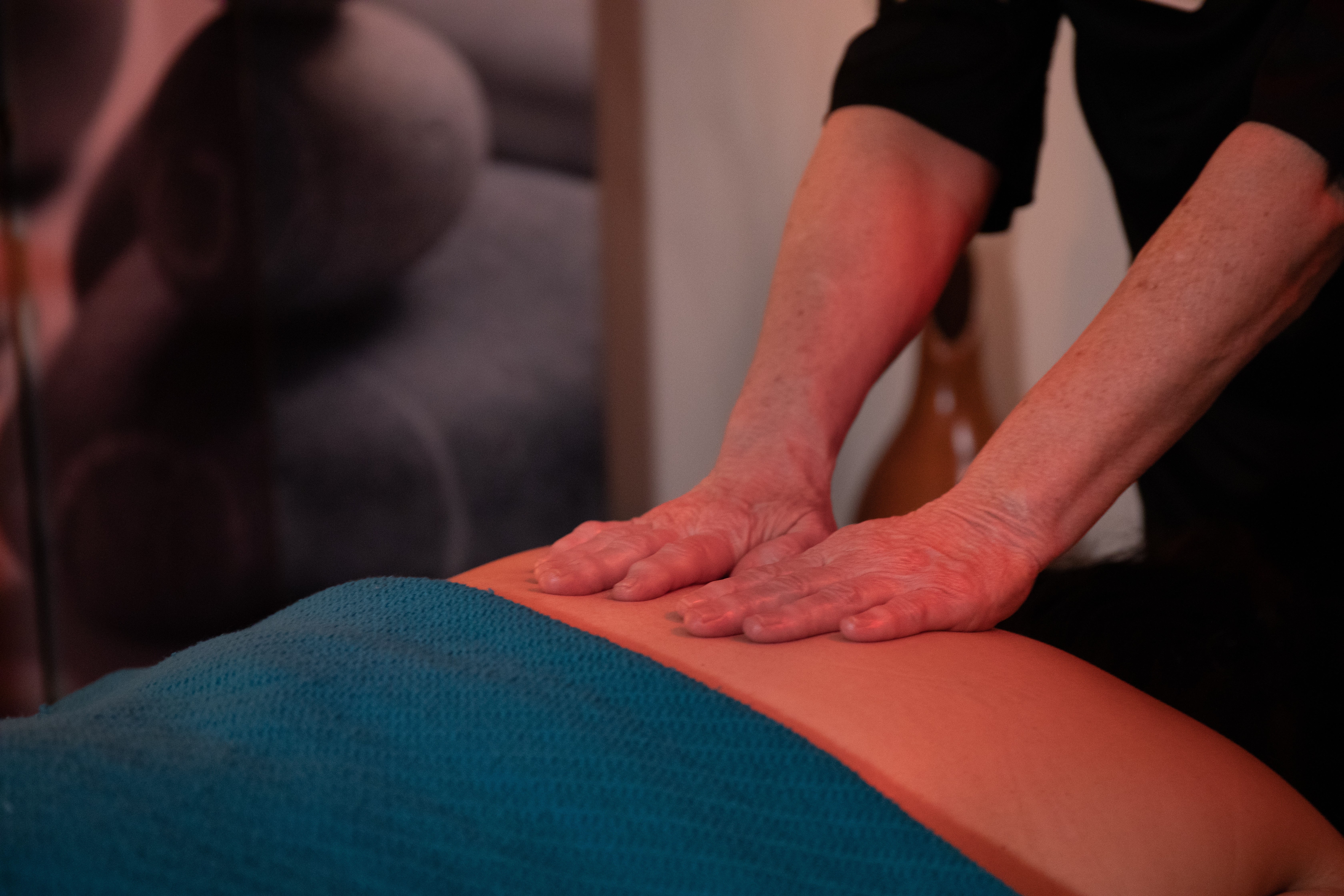 A therapist's hands press into a person's lower back under a teal blanket in a dim, warm-lit room.