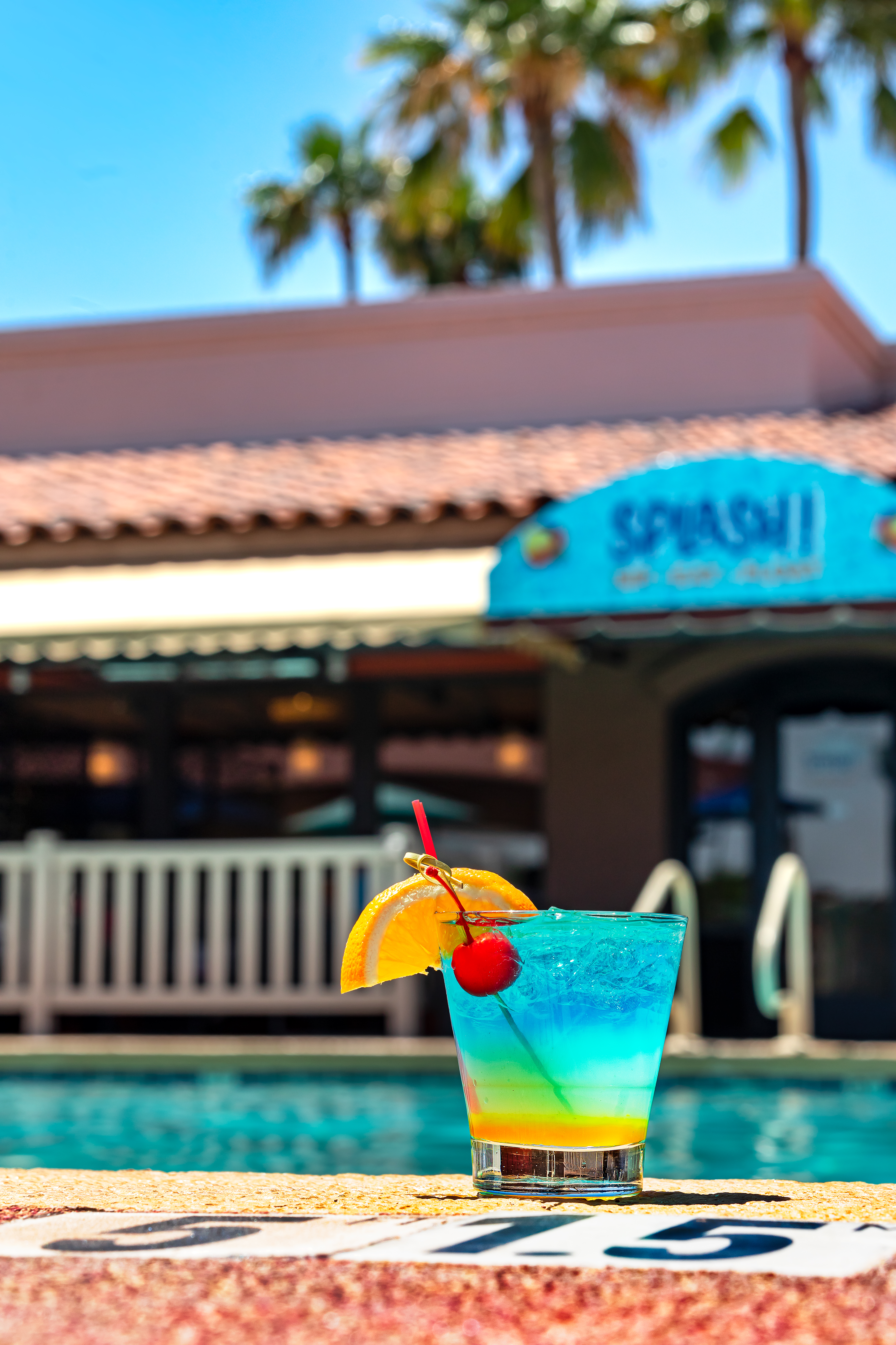A blue-and-orange cocktail with cherry and orange slice sits at a pool edge near a railing, a SPLASH! sign behind.