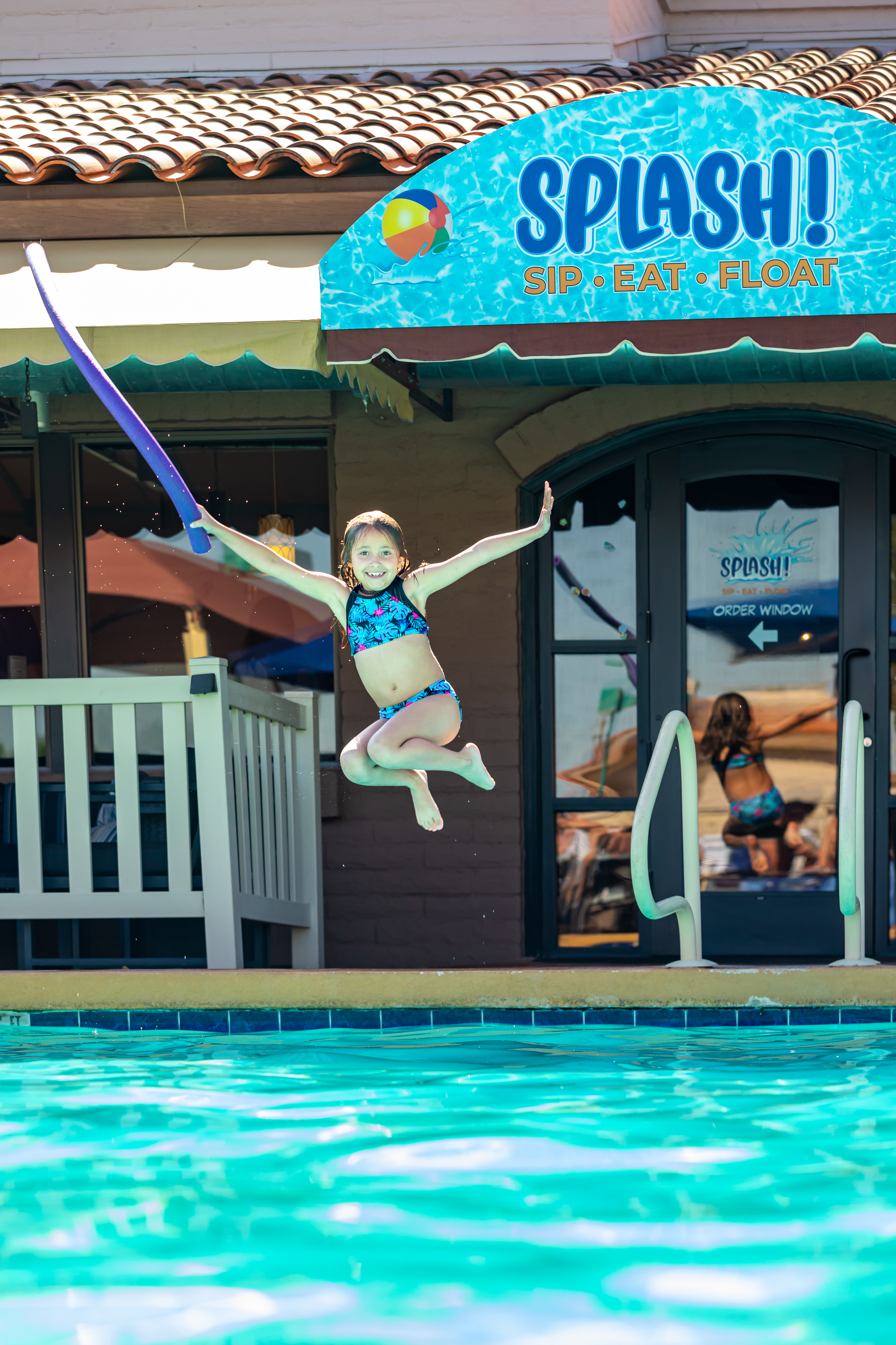 A child in a swimsuit is caught mid-jump above a turquoise pool, near a railed deck and a SPLASH! sign.