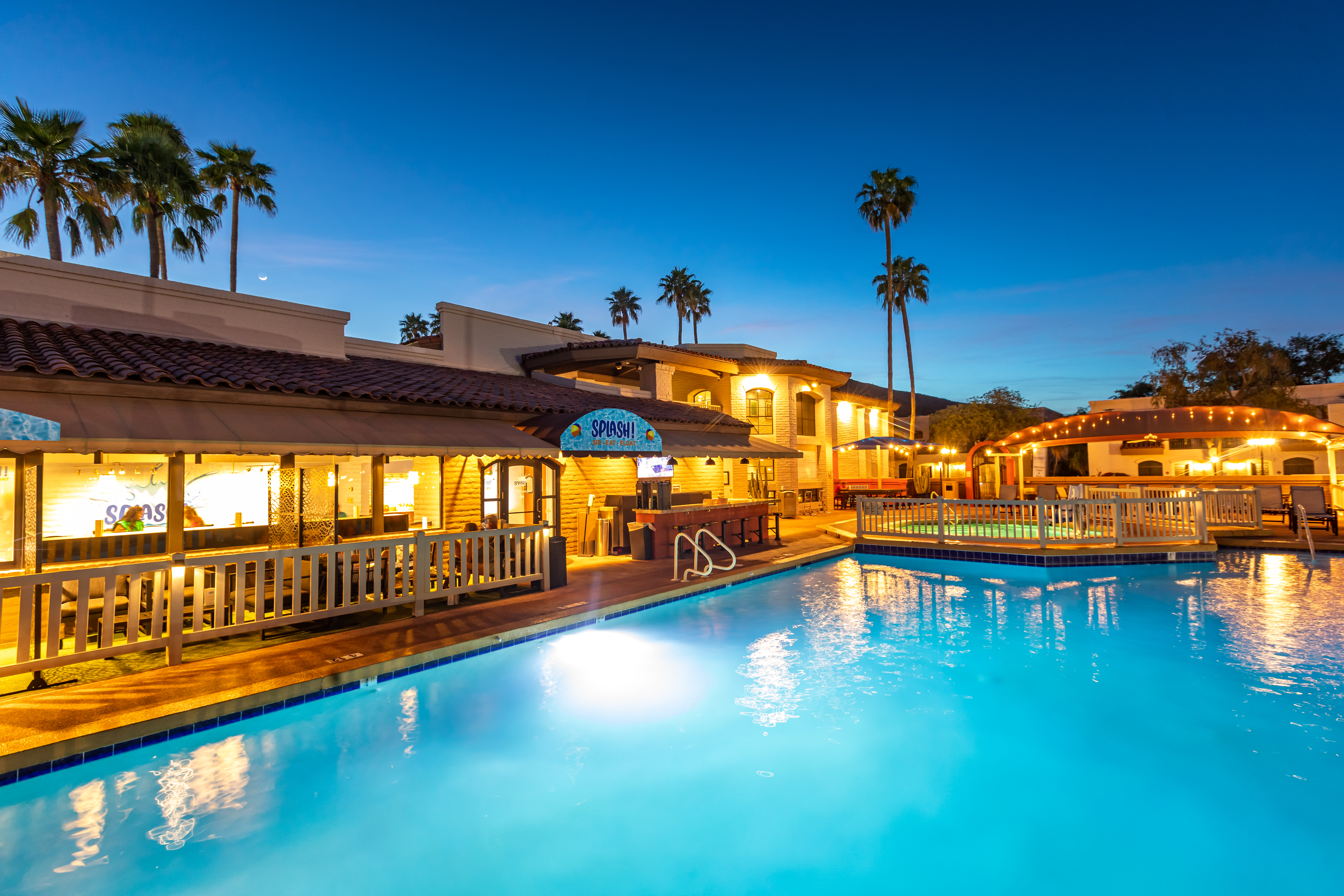 A lit blue pool sits before a low building with a red tile roof, white railings and a SPLASH! sign at dusk.