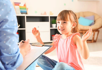 Child Therapy with psychologist with pen and clipboard as caucasian girl sits cross-legged on floor wearing jeans smiling with hands apart