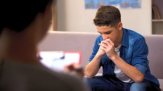 Teen Therapy at Sunstone Psychology Group with psychologist in foreground writing with clipboard and teen boy in backgroun with hands clenched thinking