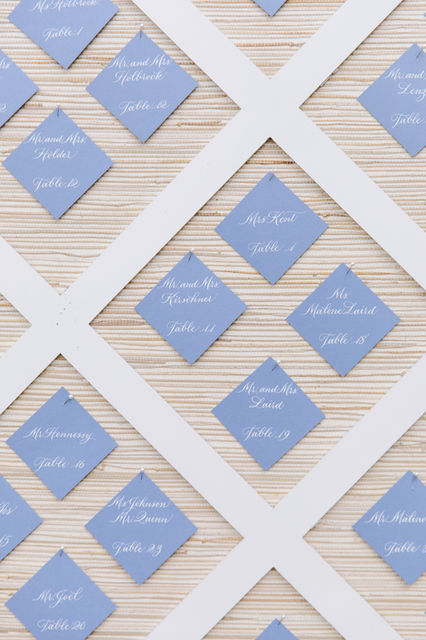 Escort cards to guide guests to their correct table.  Blue square escort cards with names and table assignments written by hand in calligraphy with white ink.  Escort cards are hung on the diagonal and displayed on a large lattice display.  