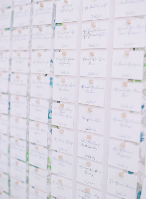 Escort cards for a wedding reception directing guests to their seating assignments. Guest names and table numbers are written by hand in blue ink on white paper in helinda rook calligraphy script. Escort cards are displayed on a large fabric colored board and affixed with gold push pins