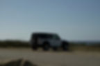 White SUV parked on a deserted coastal road in Aruba, overlooking the sea under a clear blue sky. Sparse vegetation and distant ocean view.