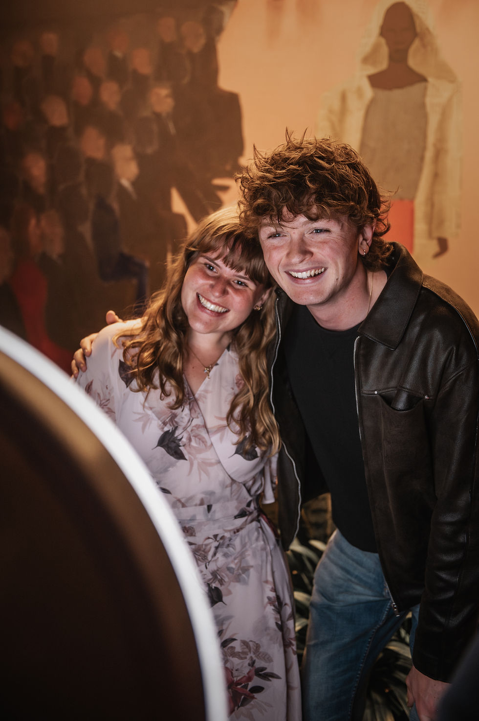 Smiling young couple pose closely indoors, woman in floral dress and man in black jacket, with blurred figures in warm background.