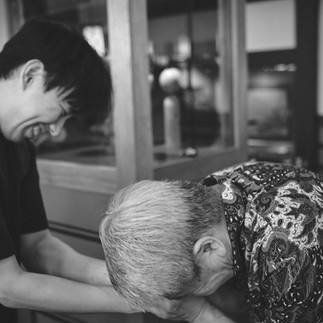 An older man bows with his forehead touching a younger man's hands.