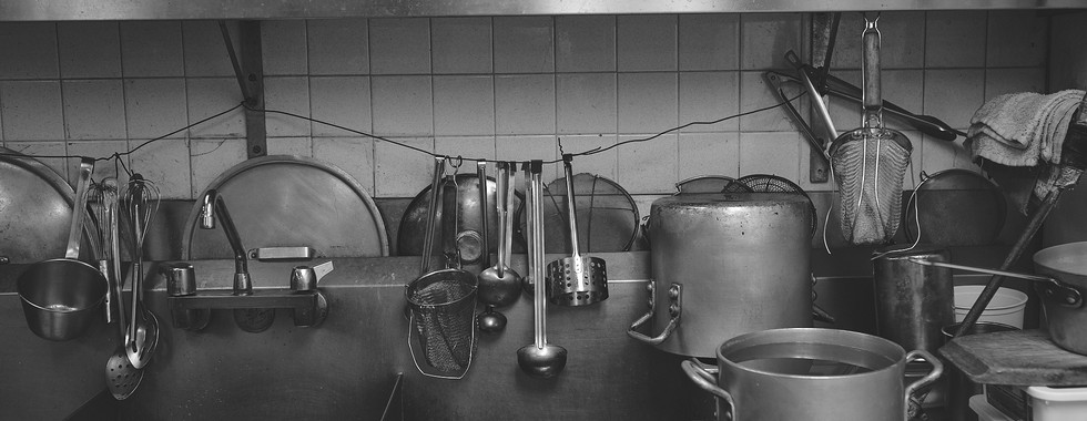 An industrial double sink with a large pot in the right hand sink. Bowls and metal containers placed on steel shelving over the sink. A long wire hangs from the shelves' support where utensils such as spoons, tongs and fine mesh strainer hang.