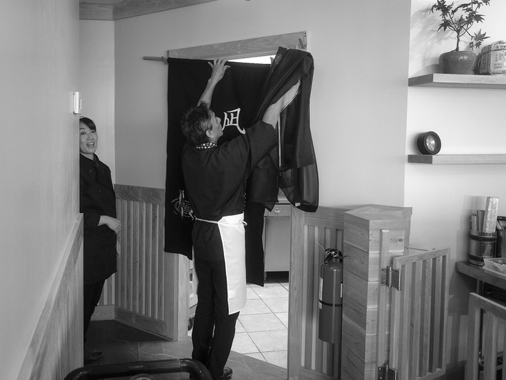 One chef replacing the noren from the dining room to the kitchen with a new noren. Another chef looks on in the background.