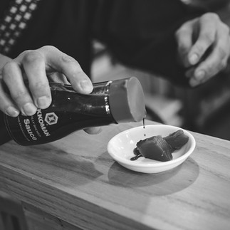 Close-up of hands pouring soy sauce over two small pieces of tuna placed in small dish.