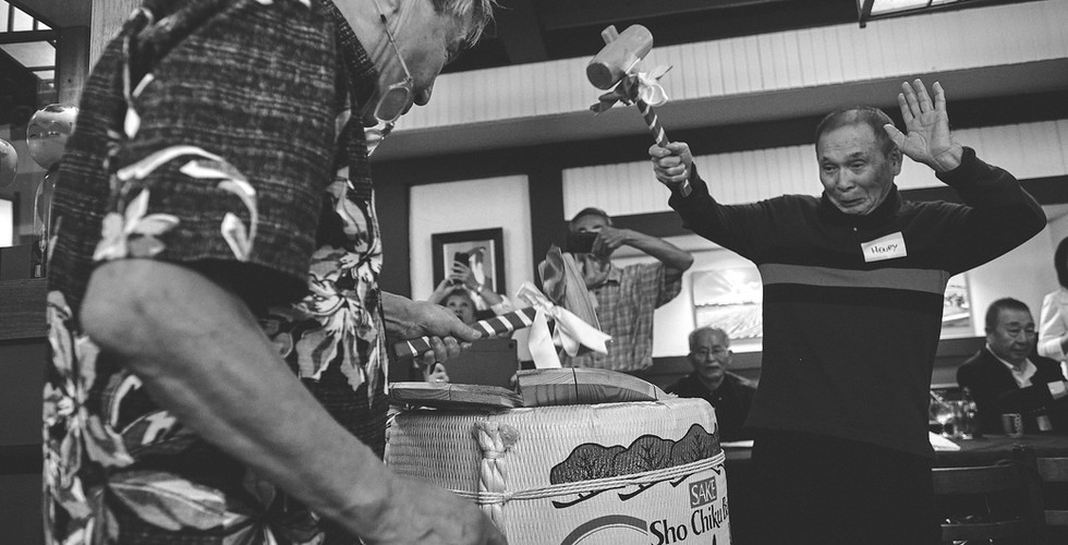 Two men joyfully open a sake barrel in a restaurant with wooden mallets, breaking the top cover, while surrounded by people taking photos. Text on the barrel reads “Sho Chiku Bai.”