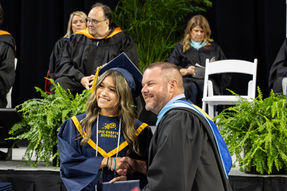 Graduates shake hands onstage at a ceremony, smiling; navy gown reads EPIC CHARTER SCHOOLS, with faculty and ferns behind.