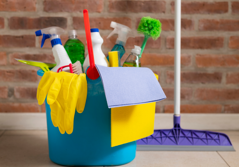 Cleaning supplies in a bucket with sprays, gloves, and mop against brick wall.