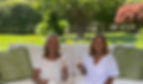 Susan and Maria, the creators of the luxury elevated dog seat, sit together on an outdoor patio couch. Both women are smiling and holding champagne glasses, dressed in white tops. The background shows a lush garden with green trees and a red flowering bush, suggesting the upscale Boca Raton setting where they developed their innovative pet furniture for small breeds.