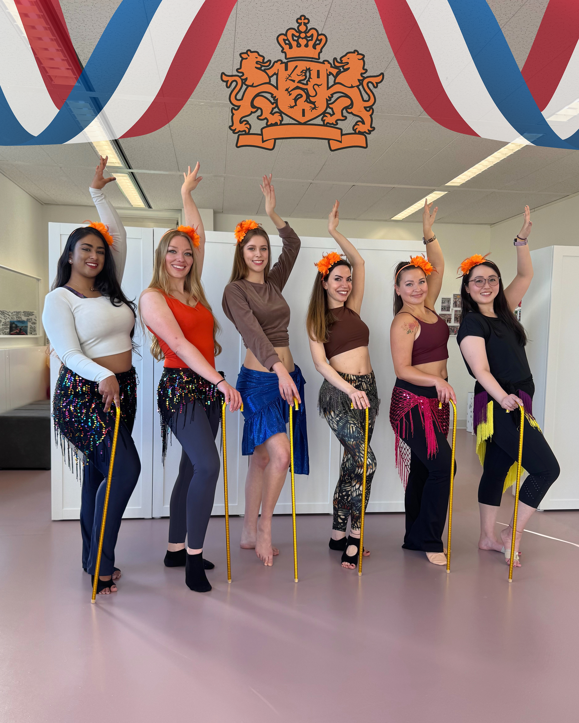 Zes buikdansstudenten op een rij met gouden stokken onder het Nederlands koninklijk wapen bij Joy Dance Lab Eindhoven